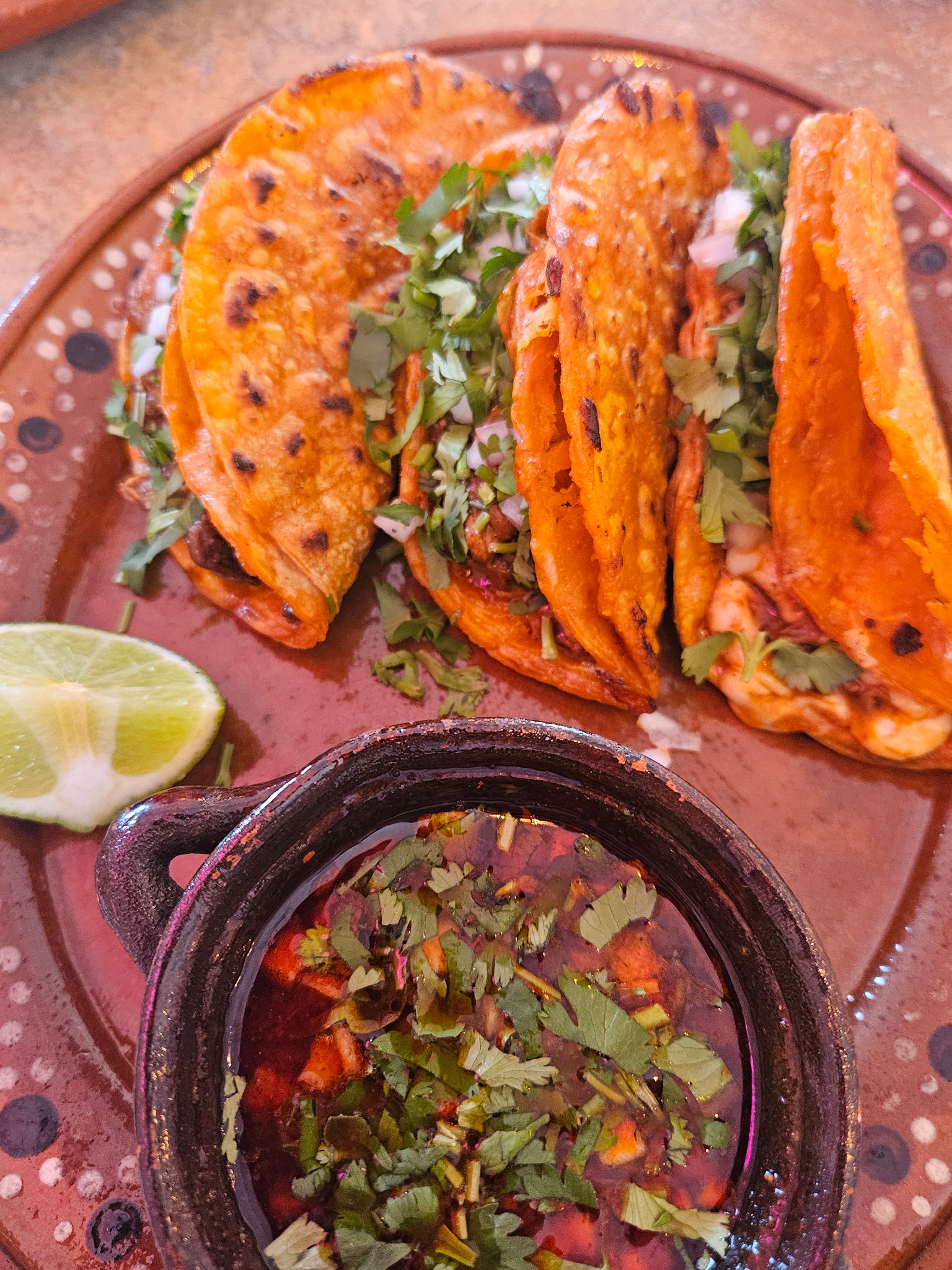 Bright-red crispy birria tacos with a cup of consomé for dipping