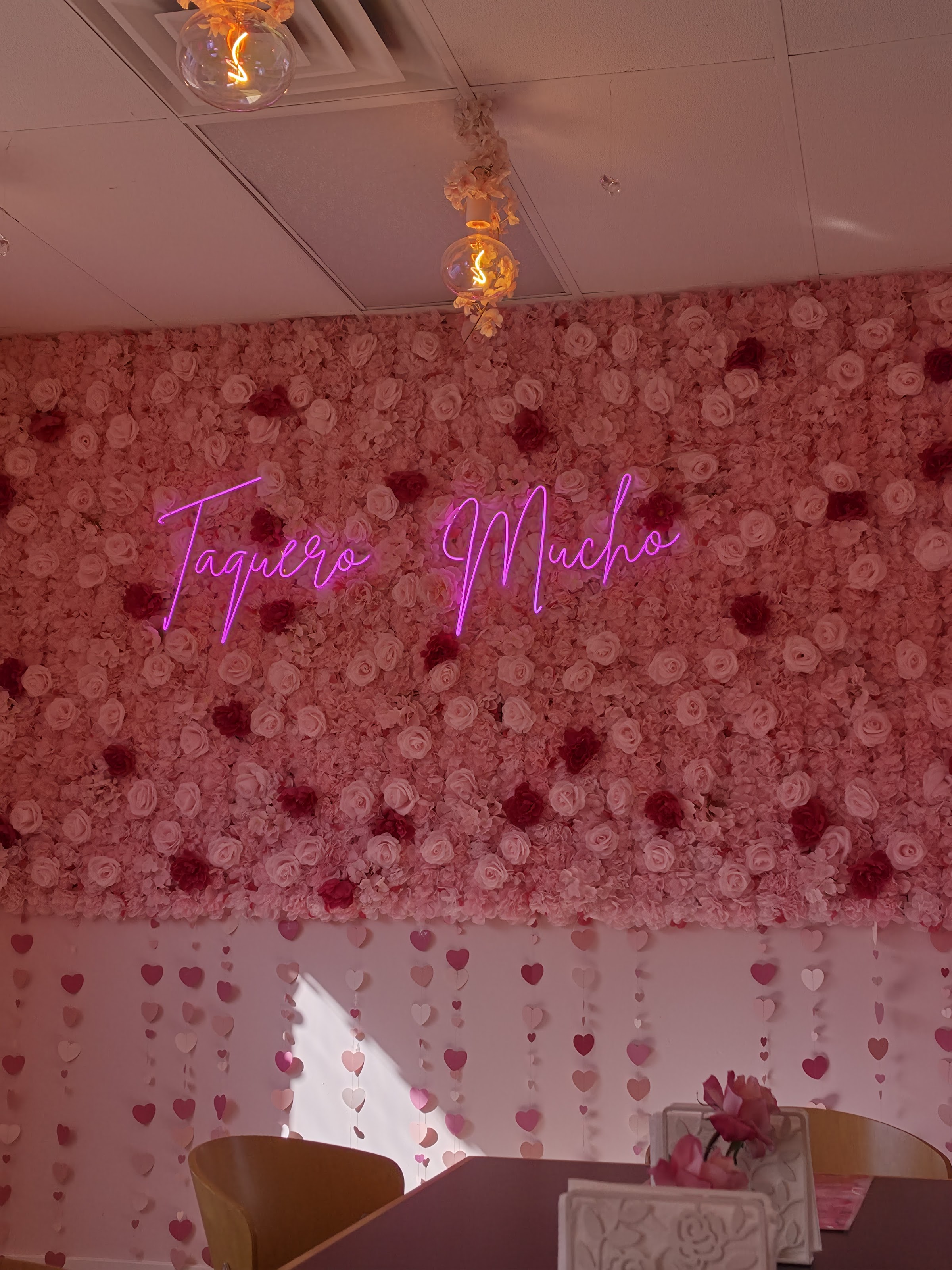 The pink-rose flower wall with the magenta 'Taquero Mucho' neon script and a hanging hearts curtain